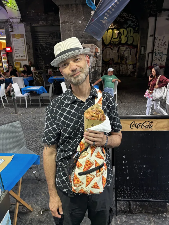 Patrick standing on a Naples street holding a wrapped cone of fried seafood, wearing a pizza-print bag