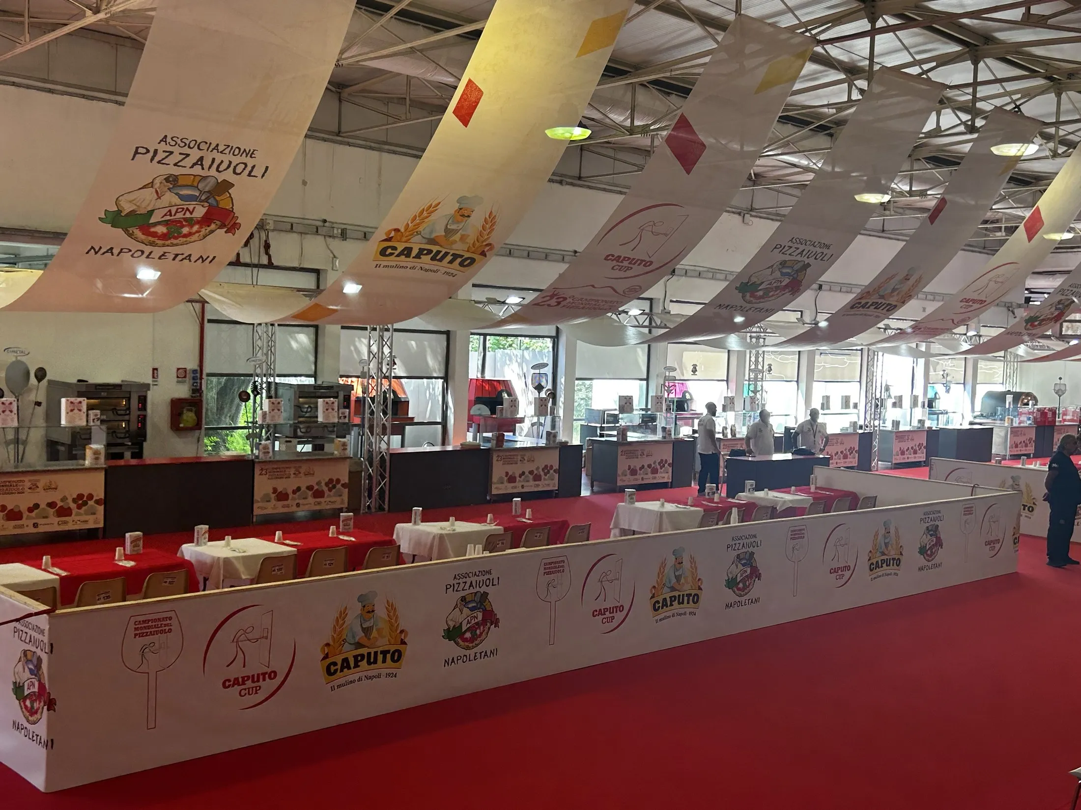 Red Caputo Cup competition floor with pizza stations, sponsor banners, ovens, and long white tables inside the Naples venue