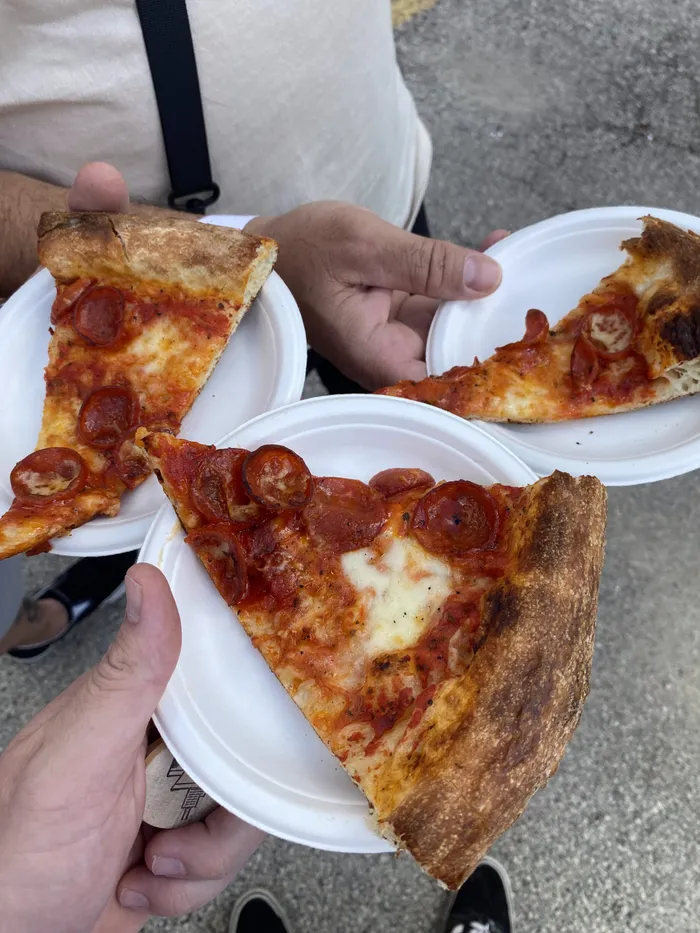 Hands holding up three thin-crust tasting slices on paper plates