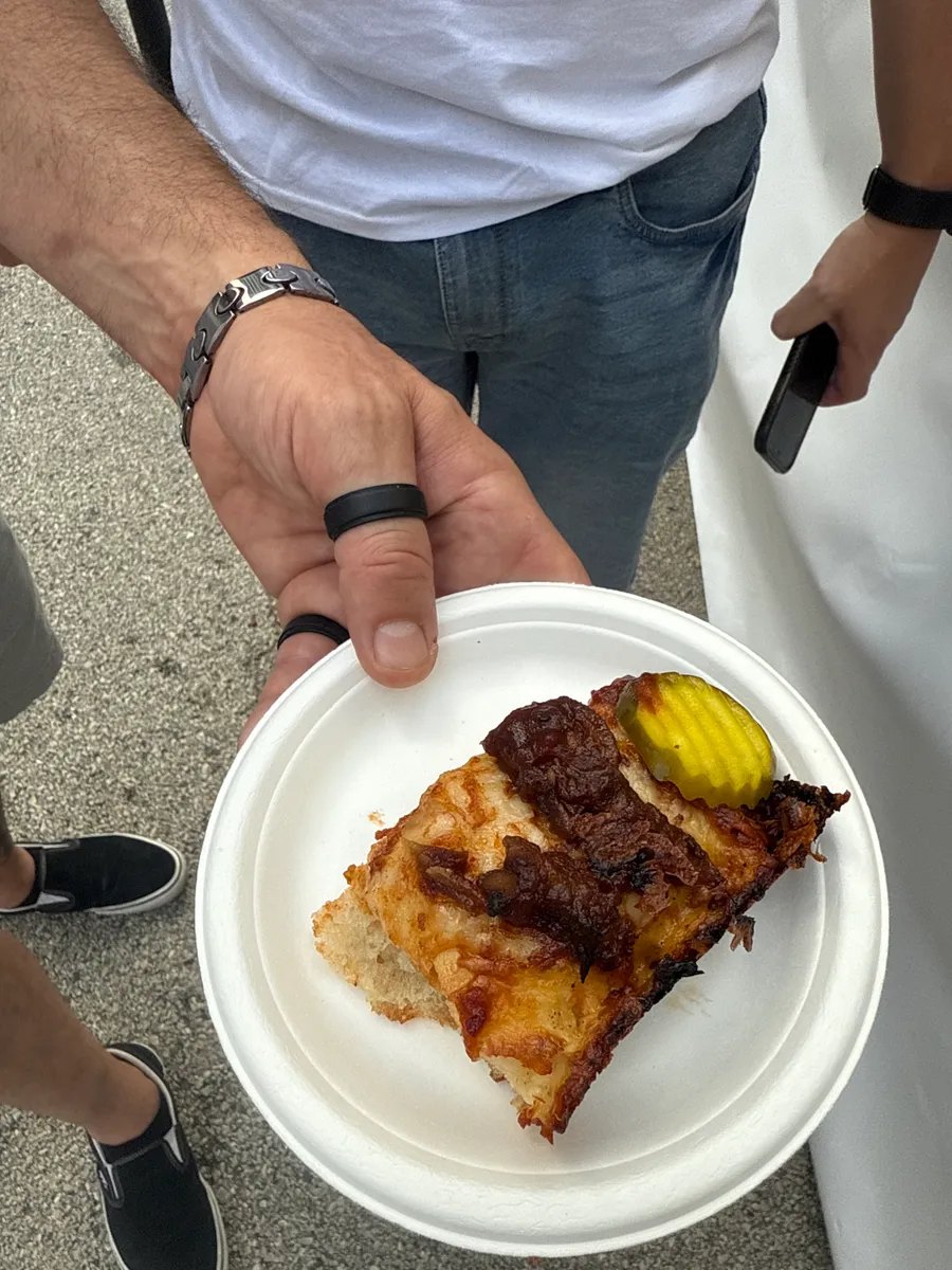 A Detroit-style tasting square topped with a pickle slice, held on a paper plate