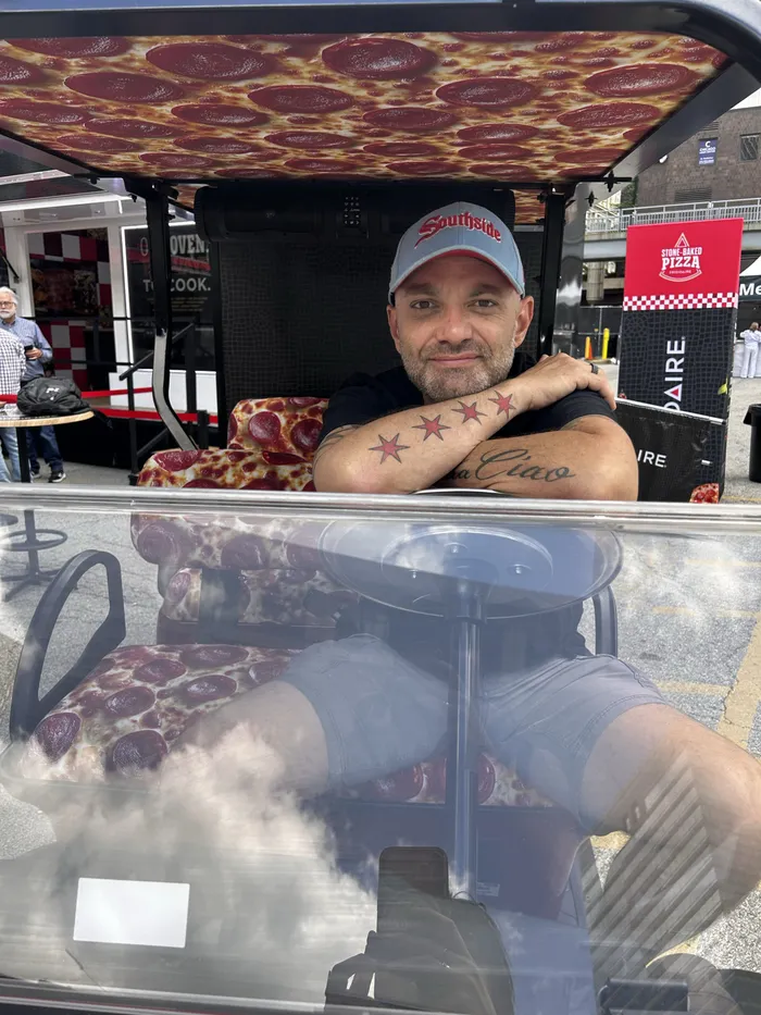 A slice scout grinning from the driver seat of a pepperoni-print golf cart at Pizza City Fest