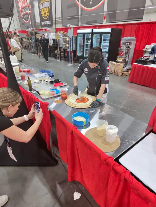 A demonstrator stretching dough at a prep table while an attendee films on a phone