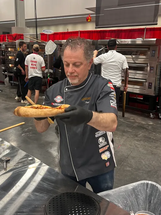 A competitor in black gloves carrying a finished pie past the competition ovens