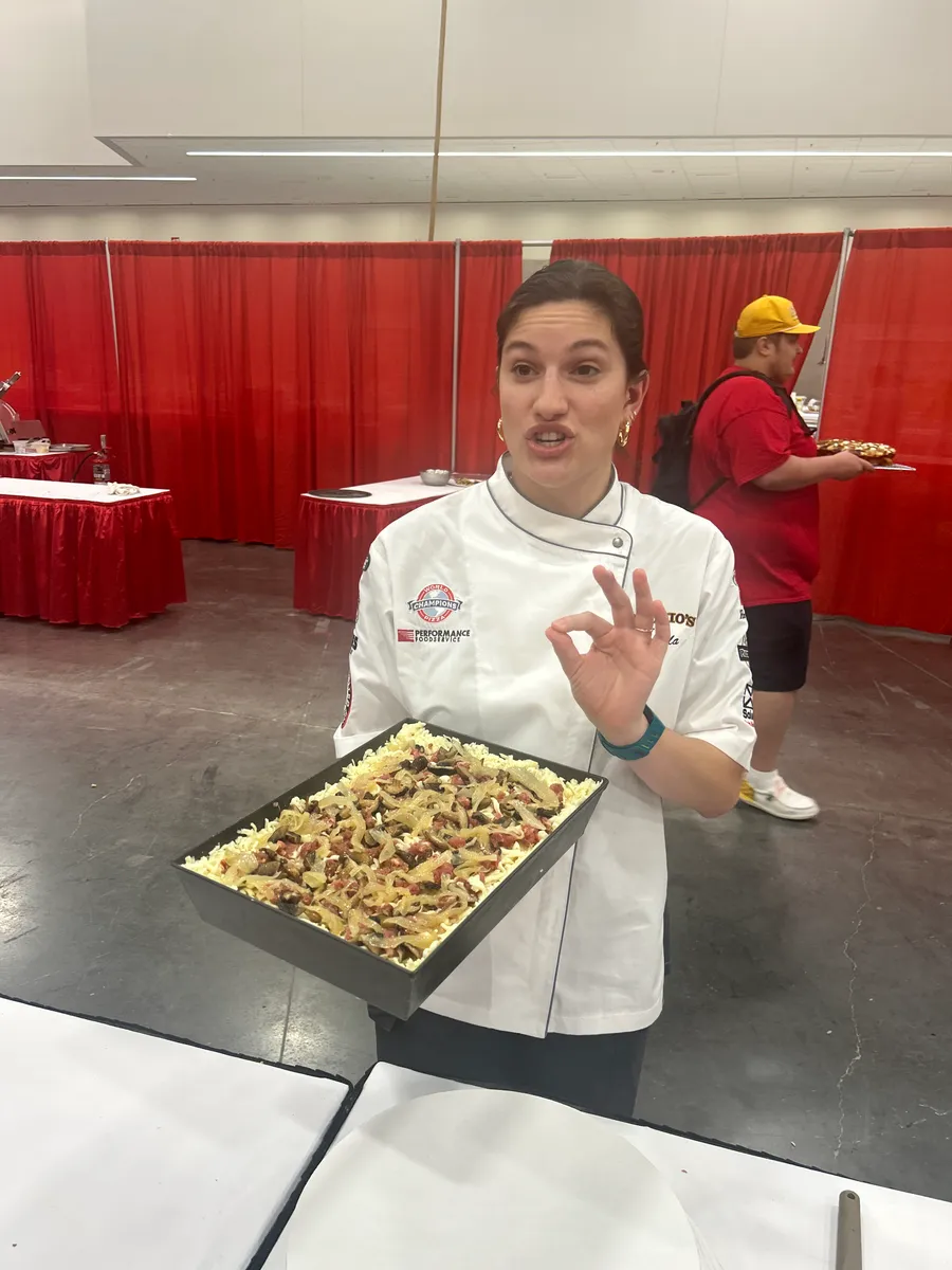 A chef in whites flashing an OK sign while presenting a sheet-pan pizza to the judges