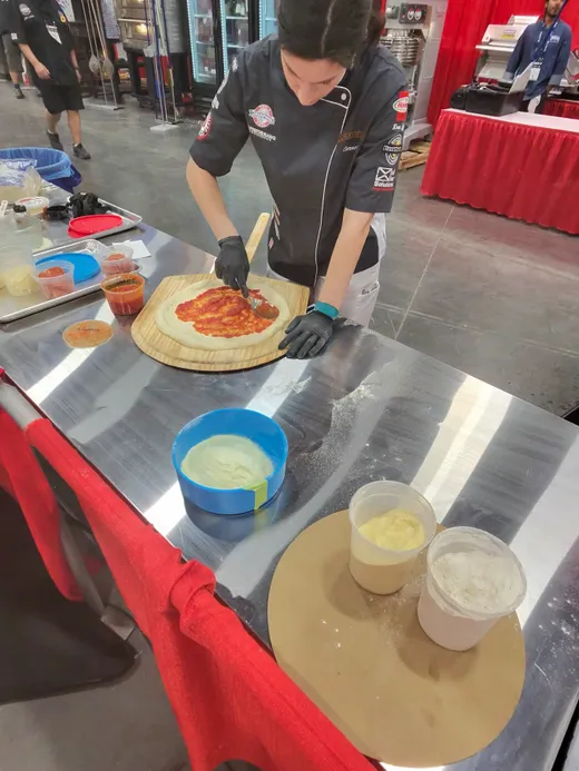 A competitor saucing a stretched dough at a prep station, sauce bowl and dough trays in front