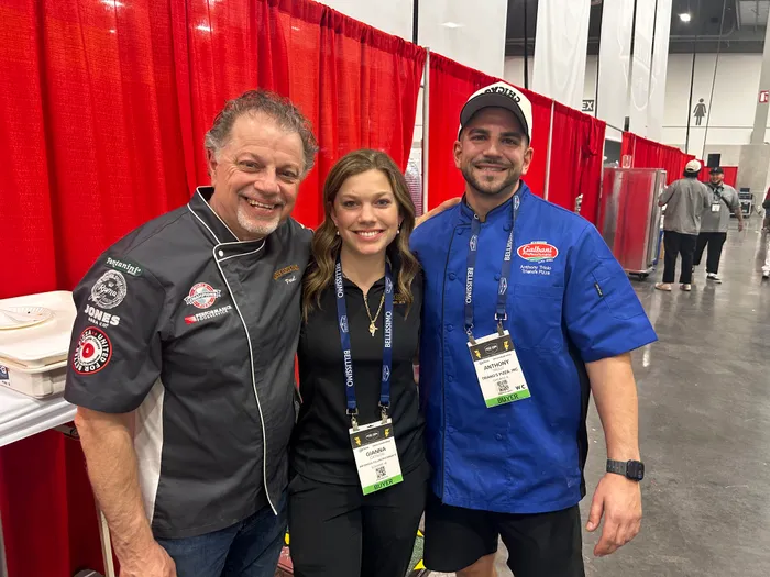 A patch-jacketed pizzaiolo, a friend, and a slice scout posing on the expo floor