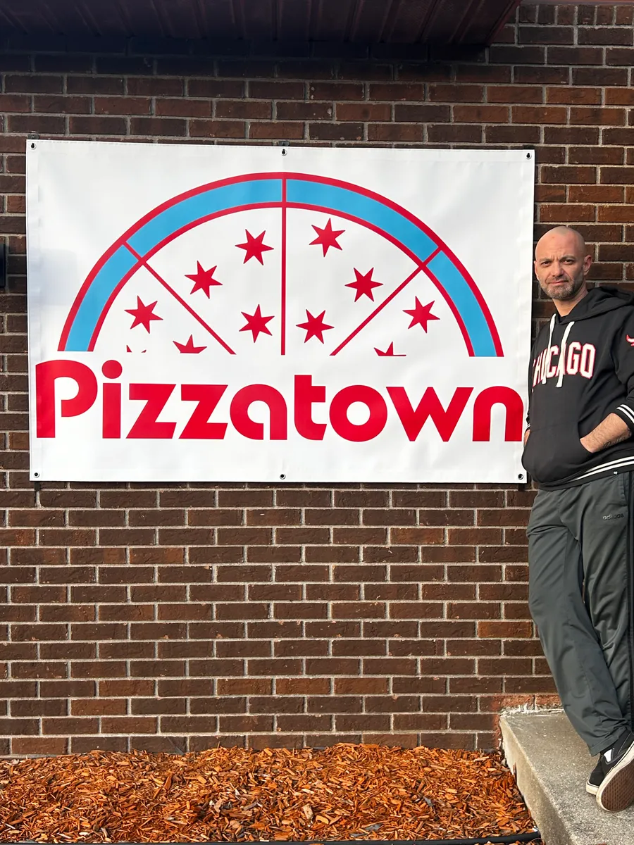 A slice scout posing beside the Pizzatown banner on a brick wall