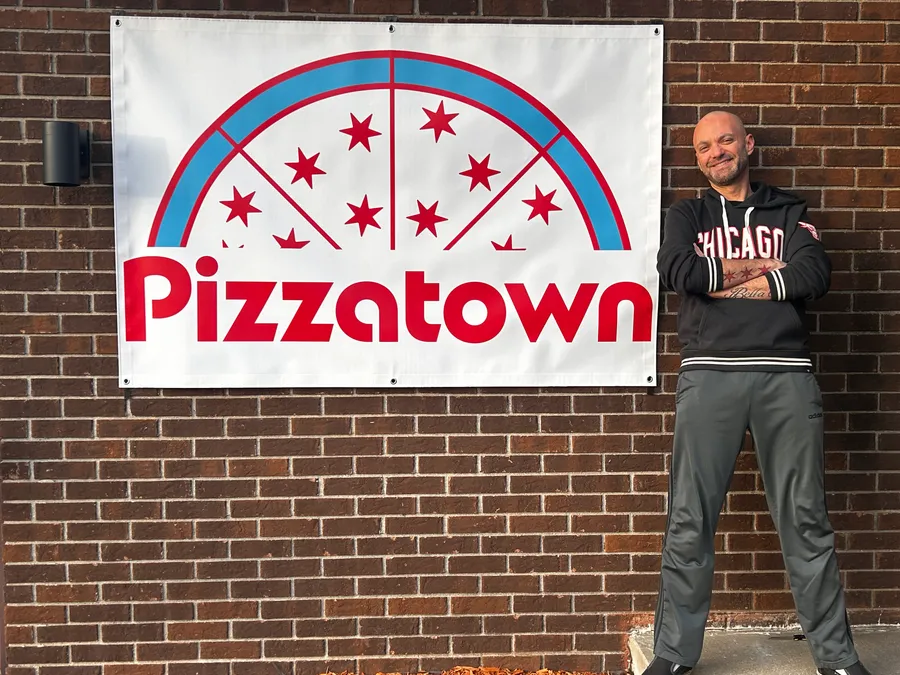 A slice scout pointing at the Pizzatown skyline-pizza banner on a brick wall