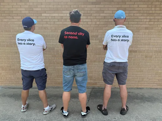 Three crew backs in a row: "Every slice has a story" and "Second city to none" shirts against a brick wall