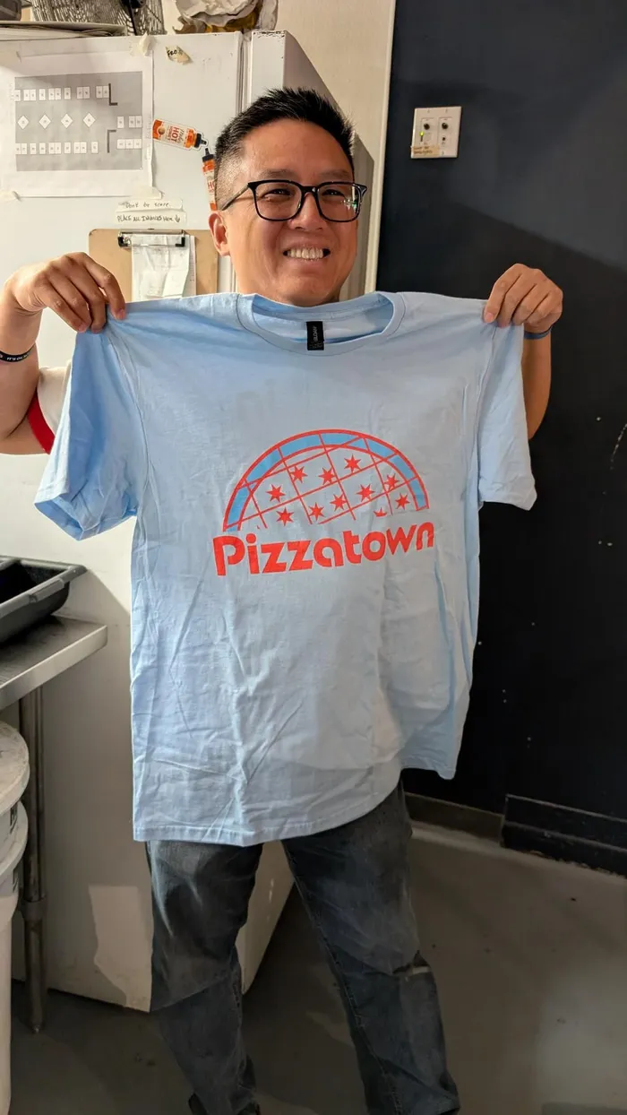 Derrick Tung of Paulie Gee's holding up a Pizzatown tee in his shop
