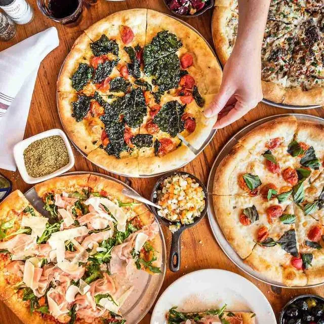 A spread of Quartino Neapolitan pizzas and small plates crowding a wood table, a hand reaching for a slice
