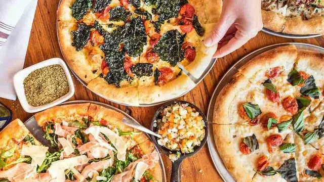 A spread of Quartino Neapolitan pizzas and small plates crowding a wood table, a hand reaching for a slice
