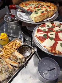 Deep dish and thin-crust pies on metal trays crowding the counter at Vero Chicago Pizza, fries and fixings alongside