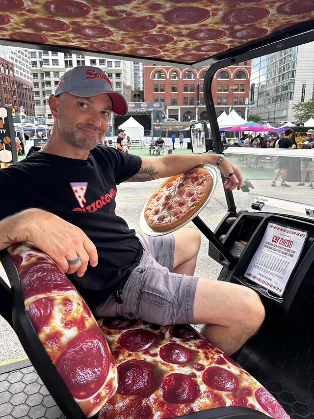 A slice scout at the wheel of a pepperoni-print golf cart at Pizza City Fest, pie in hand