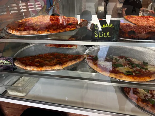 Zaza's glass slice case stacked with whole pies and a chalkboard 'cheese slice' sign